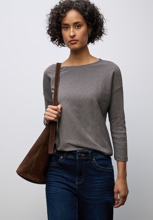 Grau gestreiftes Langarmoberteil, kombiniert mit dunkelblauen Jeans. Das Model hält eine braune Wildleder-Tasche. Einfarbiger heller Hintergrund.