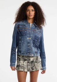 Giacca in denim con un design a strappi e dettagli splatter, due tasche sul petto e una vestibilità cropped. Abbinata a una gonna a pieghe con motivo mimetico.