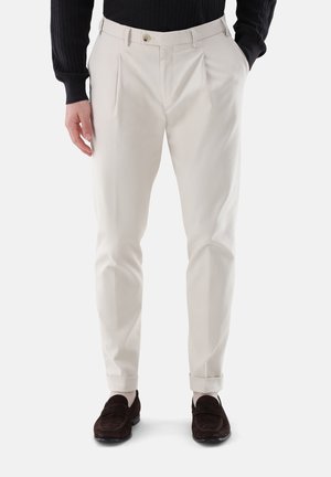 Mann trägt beige plissierte Hose mit Umschlägen, einen schwarzen Pullover, weiße Socken und dunkelbraune Loafers, steht mit Händen in den Taschen.