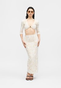 Γυναίκα που φοράει εκρού κορυφή από δαντέλα με μανίκια τριών τετάρτων και ταιριαστό μακρύ ψηλόμεσο φούστα, μαύρες ανοικτές γόβες και κοκκάλινα πορτοκαλί γυαλιά ηλίου.