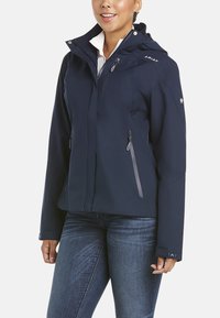 Veste en softshell bleu marine avec une coupe ajustée, capuche, poches zippées et accents gris sur les zips et le logo. Légère avec une texture lisse.