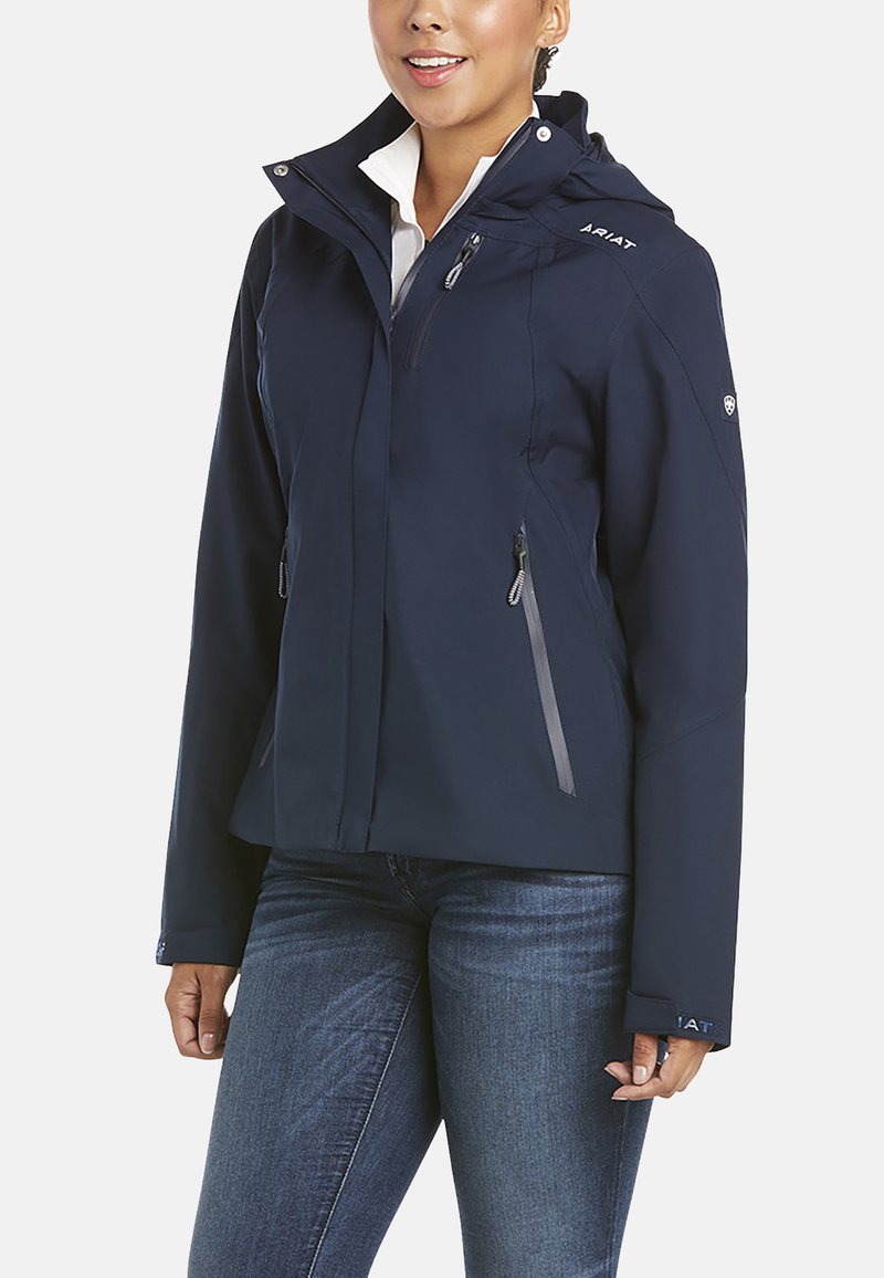 Veste en softshell bleu marine avec une coupe ajustée, capuche, poches zippées et accents gris sur les zips et le logo. Légère avec une texture lisse.