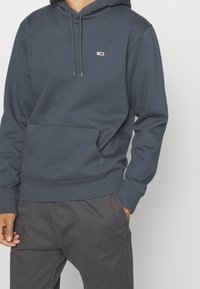 Hombre con sudadera con capucha gris oscuro con bolsillo frontal y pequeño logo rectangular en el pecho, combinado con pantalones grises, mano en el bolsillo.