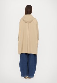 Giacca beige con cappuccio e vestibilità ampia, con una fessura sul retro, abbinata a pantaloni in denim blu scuro a gamba larga e scarpe chiare.