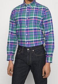 Green and purple checkered button-up shirt with white buttons, featuring a classic collar. Worn with dark denim jeans.