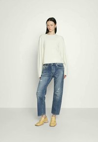 Frau mit glatten, dunklen Haaren, die einen weißen Pullover, ausgewaschene blaue Jeans und gelbe Slipper trägt, steht vor einem schlichten hellen Hintergrund.