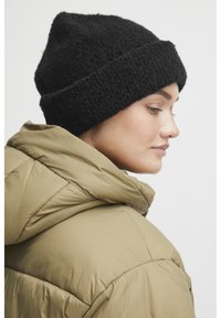 Svart teksturert strikket lue med brett, sammen med en pufferjakke i olivengrønn, vist fra sideprofil mot en nøytral bakgrunn.