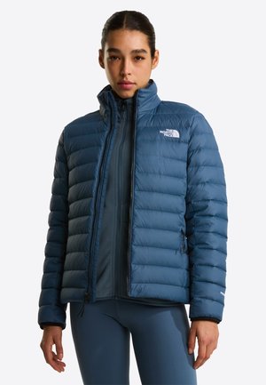 Kobieta w niebieskiej puchowej kurtce z zamkiem z przodu i wysokim kołnierzem, stojąca w zrelaksowanej pozie na jednolitym tle.