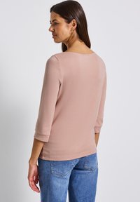 Frau mit braunem Haar, die ein hellrosa Oberteil mit drei Viertelärmeln und blaue Jeans trägt. Sie ist leicht zur Seite gedreht vor einem schlichten Hintergrund.