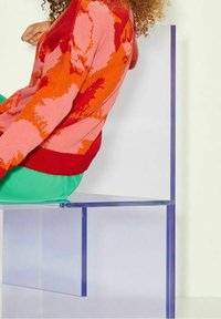 A transparent acrylic chair with a minimalist design. The seated person wears a patterned red and pink sweater and green pants.