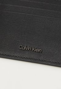 Svart teksturert skinnkortholder med flere kortspor og preget Calvin Klein-logo, med subtile sømmer.