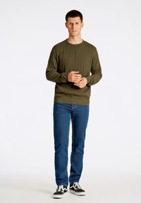 Maglione verde oliva a maglia cable con scollo rotondo, abbinato a jeans denim blu e sneaker in bianco e nero. Texture liscia, vestibilità casual.