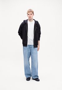 Čierna mikina na zips s červenými akcentmi, biele tričko, svetlomodré široké džínsy a čierne topánky, ktoré nosí stojaci model.