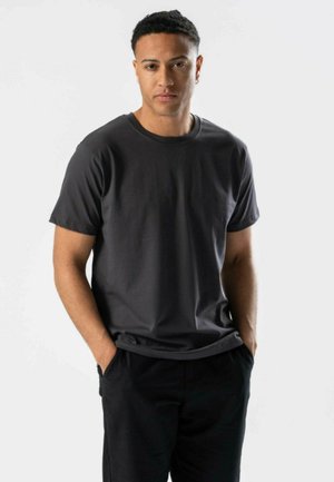 Ung mand med kort krøllet hår står med hænderne i lommerne iført en ensfarvet mørkegrå t-shirt og sorte bukser mod en hvid baggrund.