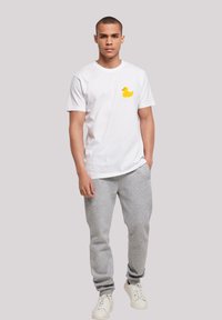 White cotton t-shirt featuring a yellow rubber duck graphic on the left chest, paired with gray sweatpants and white sneakers.