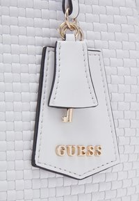 Texture en cuir tissé blanc avec une étiquette en cuir blanc présentant une petite clé dorée et des lettres dorées "GUESS".