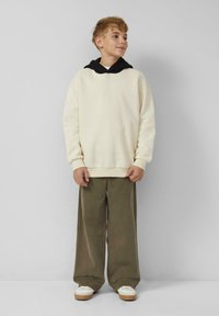 Ein Junge mit kurzem, hellbraunem Haar, der einen cremefarbenen Hoodie mit schwarzer Kapuze, olivfarbenen weit geschnittenen Hosen und weißen Turnschuhen trägt, steht an einer schlichten Wand.