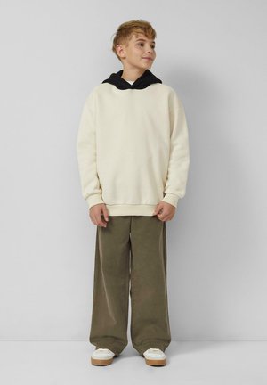 Ein Junge mit kurzem, hellbraunem Haar, der einen cremefarbenen Hoodie mit schwarzer Kapuze, olivfarbenen weit geschnittenen Hosen und weißen Turnschuhen trägt, steht an einer schlichten Wand.