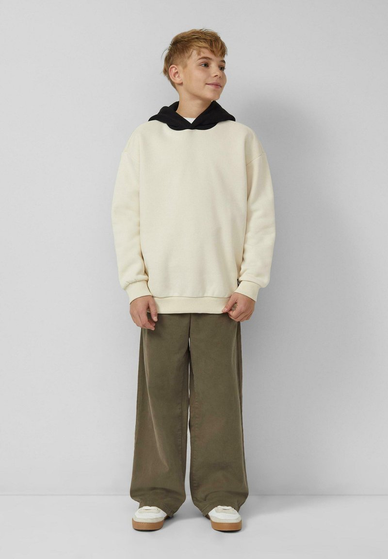 Ein Junge mit kurzem, hellbraunem Haar, der einen cremefarbenen Hoodie mit schwarzer Kapuze, olivfarbenen weit geschnittenen Hosen und weißen Turnschuhen trägt, steht an einer schlichten Wand.