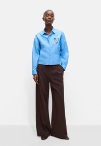 Jasnoniebieska koszula na guziki z sercowym wzorem na lewej piersi, w połączeniu z brązowymi szerokimi spodniami, na białym tle.