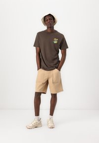 Braunes Baumwoll-T-Shirt mit grafischem Design, kombiniert mit beigen Shorts mit Fronttaschen. Das Modell trägt beige Sneaker und einen cremefarbenen Bucket-Hut.