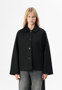 En lys blond kvinne iført en løs svart knappeløs jakke med vide ermer og falmede svarte jeans, som står mot en hvit bakgrunn.
