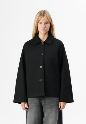Blonde vrouw in een losse zwarte button-up jas met wijde mouwen en vervaagde zwarte jeans, staande tegen een witte achtergrond.
