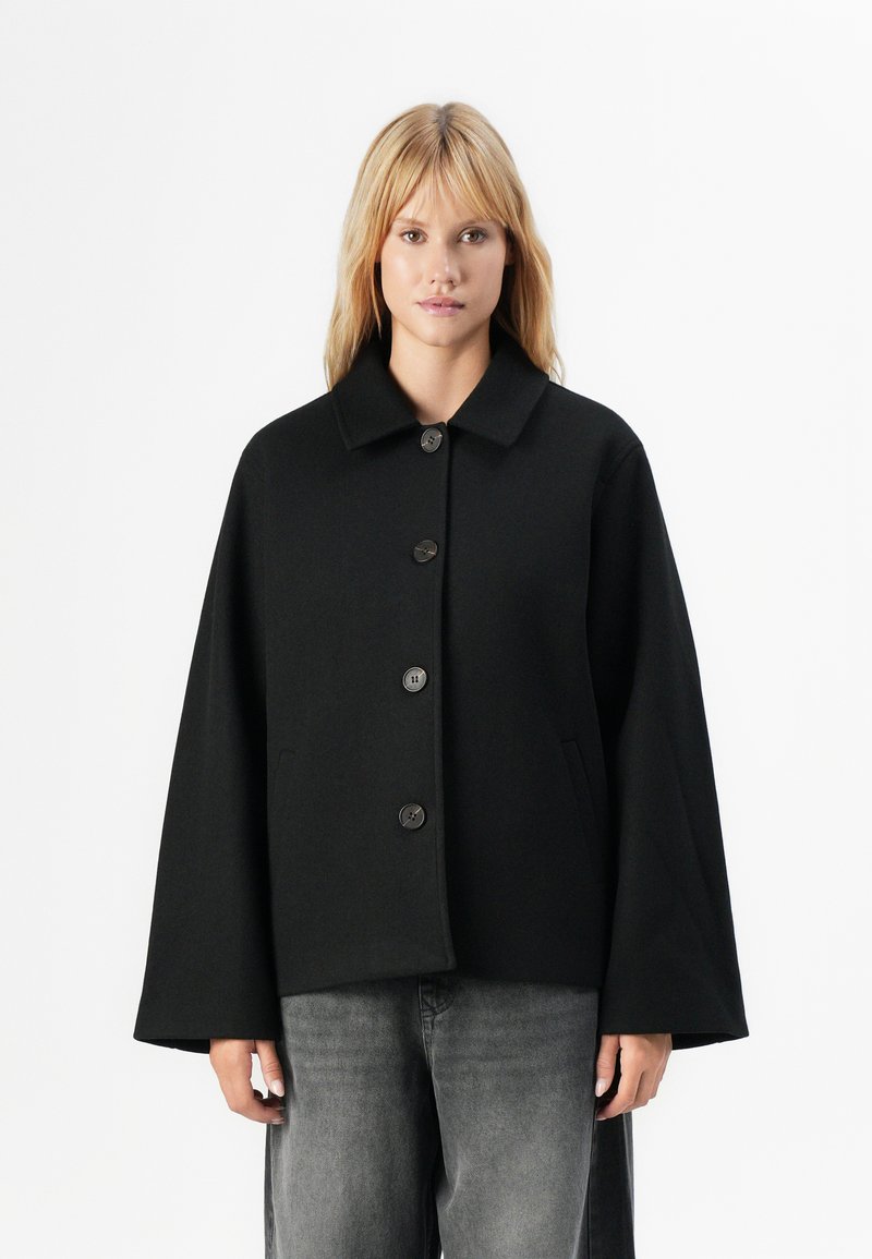 En lys blond kvinne iført en løs svart knappeløs jakke med vide ermer og falmede svarte jeans, som står mot en hvit bakgrunn.