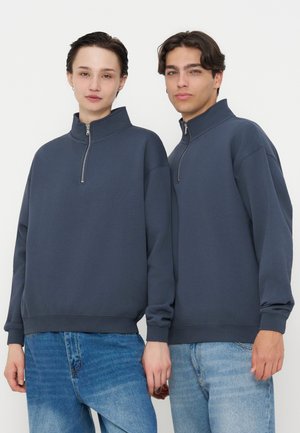 Deux personnes portant des sweats à capuche gris foncé zippés assortis et des jeans bleus, debout côte à côte sur un fond blanc.