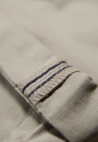 Beige kangasta, jossa on tekstuuri, sisältää kontrastoivia laivastonsinisiä ompeleita ja käänteisen hihansuiden yksityiskohdan. Siistit reunat ja sileä pinta näkyvissä.