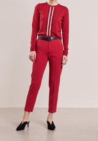 Punainen villatakki, jossa on röyhelöinen etuosa ja napit, yhdistettynä muotoiltuihin punaisiin housuihin. Musta vyö ja mustat korkokengät täydentävät asukokonaisuuden.