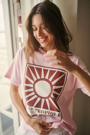 Jeune femme souriante, les yeux fermés, portant un t-shirt rose avec un motif de soleil rouge près d'une fenêtre à l'intérieur.