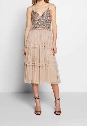 Vrouw draagt een mouwloze jurk met een roségouden pailletten lijfje en een beige gelaagde tule midi-rok, gecombineerd met zwarte sandalen met bandjes en hak.