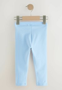 Hellblaue weiche Baumwoll-Babyleggings an einem Holzbügel mit Metallklammern vor einem schlichten weißen Hintergrund aufgehängt.