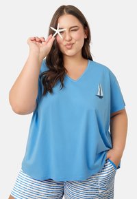 Niebieska koszulka z dekoltem w kształcie litery V z miękkiej tkaniny, z motywem żaglówki. Połączona z białymi szortami ozdobionymi niebieskimi paskami.