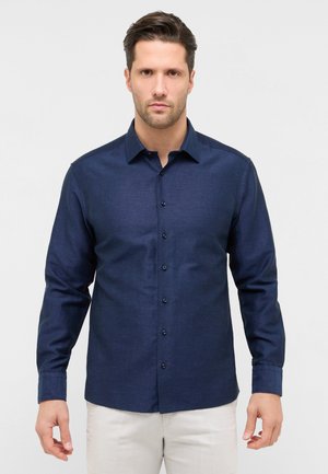 Homme portant une chemise bleu marine à manches longues boutonnée et un pantalon clair, debout devant un fond uni clair.
