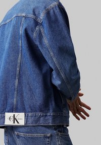 Džínová bunda ve středně modré barvě s volným střihem, viditelným švem a značkovou nášivkou na zadním lemu. Měkká textura a neformální design.