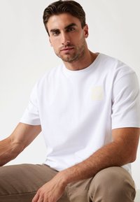 Hvid bomulds-t-shirt med korte ærmer og en rund halsudskæring, der har et kvadratisk beige mærke med "GUESS" i midten.