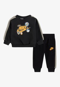Crni set dukserice i hlača s narančastim Nike logotipom. Ima sive akcentne detalje i razne zakrpe na dukserici. Mekana, rastezljiva tkanina.