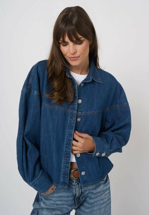 Kobieta o długich brązowych włosach, ubrana w luźną niebieską dżinsową kurtkę na białą koszulę i niebieskie jeansy, stojąca na jasnym, jednolitym tle.