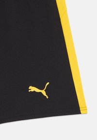 Svarte treningsshorts med en gul side stripe og en liten gul Puma-logo på venstre ben. Laget av teksturert, lett materiale.