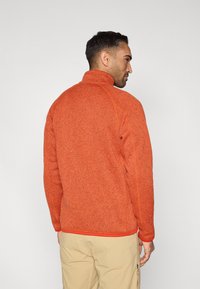 Φλις πουλόβερ σε έντονο πορτοκαλί με ψηλό γιακά, υφασμάτινη υφή και αντίθετες κόκκινες λεπτομέρειες στο τελείωμα και τις μανσέτες.