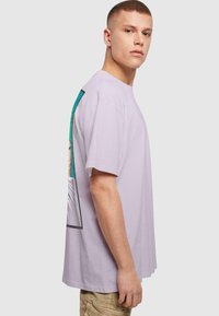 Camiseta de algodón de color púrpura claro con un gran estampado gráfico en el lado izquierdo con un diseño en verde y azul. Mangas cortas y cuello redondo.