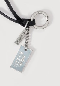 Μπρελόκ με μαύρο λουρί, ασημένιο δαχτυλίδι, αλυσίδα και δύο μεταλλικές πλακέτες με χαραγμένα τα "Calvin Klein" και "New York" σε λευκό φόντο.