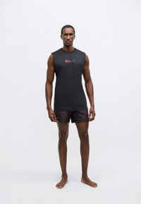 Črna majica brez rokavov z rdečim logotipom "HUGO", združena s črnimi kratkimi hlačami z diskretnim blagovnim znakom. Gladek material in atletski kroj.