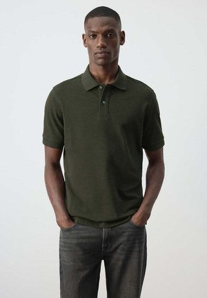 Mann mit kurzen Haaren, der ein dunkelgrünes Poloshirt mit kurzen Ärmeln und dunkle Jeans trägt, steht mit den Händen in den Taschen vor einem einfarbigen Hintergrund.