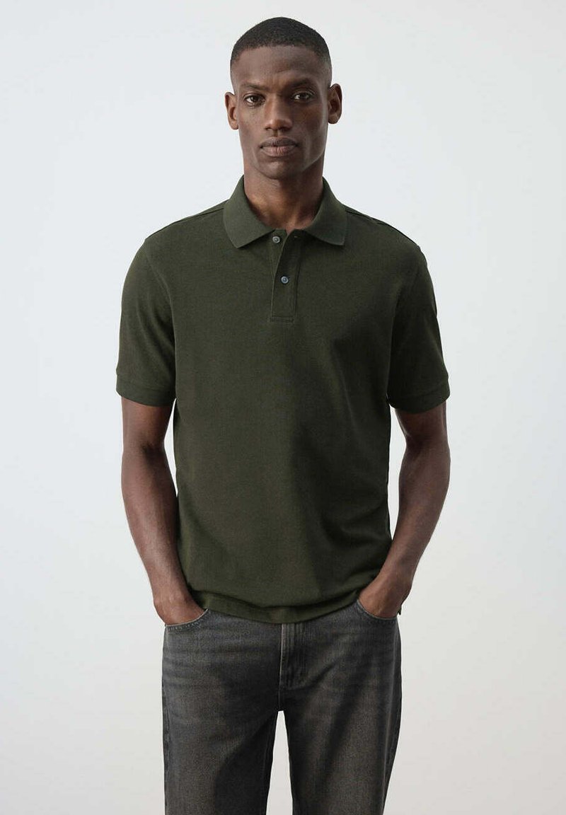Homme aux cheveux courts portant un polo vert foncé à manches courtes et un jean foncé, debout les mains dans les poches devant un fond uni.