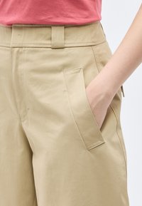 Άτομο που φοράει μπεζ παντελόνι με το χέρι στην τσέπη της δεξιάς πλευράς και ροζ πουκάμισο σολομού μέσα από το παντελόνι.