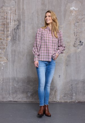 Šypsanti moteris su ilgais blondiniais plaukais, vilkinti rožine languota palaidine, šviesiai mėlynomis aptemptomis džinsais ir rudos spalvos auliniais, stovinti prie tekstūruotos sienos.