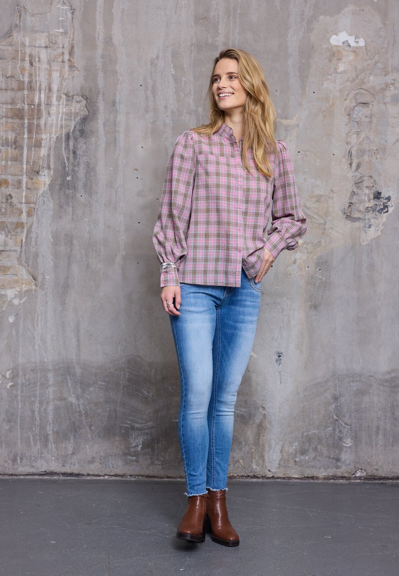 Femme souriante aux longs cheveux blonds portant un chemisier à carreaux rose, un jean skinny bleu clair et des bottines marron, debout près d'un mur texturé.